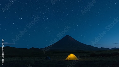 A yellow tent stands under a starry sky in a Kamchatka campsite. The silhouette of Vilyuchik volcano, shaped like a cone, rises in the dark blue night. photo