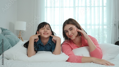 Happy asian daughter and smart mom lie on bed while looking at camera. Energetic family spend time and prepare to sleep together. Cute girl and mom smile to camera show love and affection. Pedagogy.