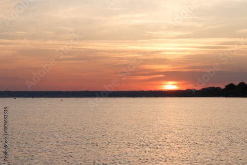 A tranquil sunrise with warm orange and golden hues reflects on the calm water, as a distant shoreline and trees create a serene silhouette.