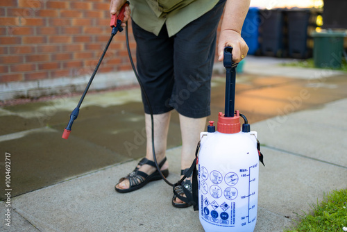 Woman spraying gray concrete patio paving slabs with liquid weed killer from pressurized plastic tank and handheld nozzle, pumping tank photo