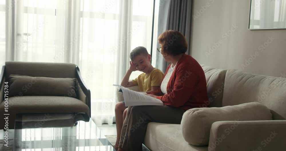 Side view. Happy family relationships. Adorable redhead little kid girl smiles, expressing happiness spending weekend with her loving grandmother at home, enjoying reading fairy tales together. Side view. Happy family relationships. Adorable redhead little kid girl smiles, expressing happiness spending weekend with her loving grandmother at home, enjoying reading fairy tales together.