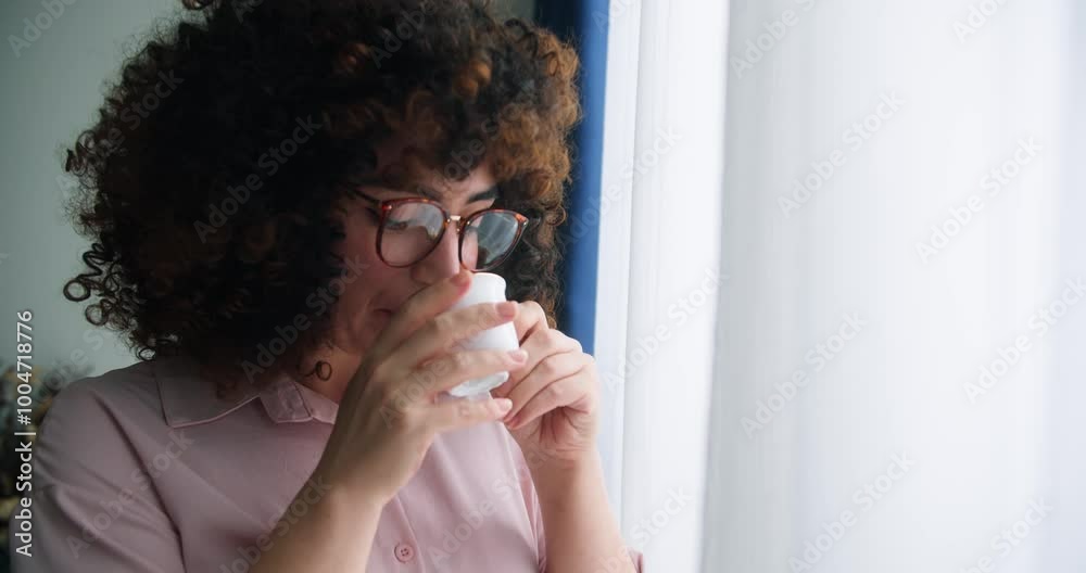 Happy positive woman with curly hair wearing glasses holding cup enjoy favorite tea drink morning invigorating coffee beverage while, the sun shines on her from the big window. Happy positive woman with curly hair wearing glasses holding cup enjoy favorite tea drink morning invigorating coffee beverage while, the sun shines on her from the big window.