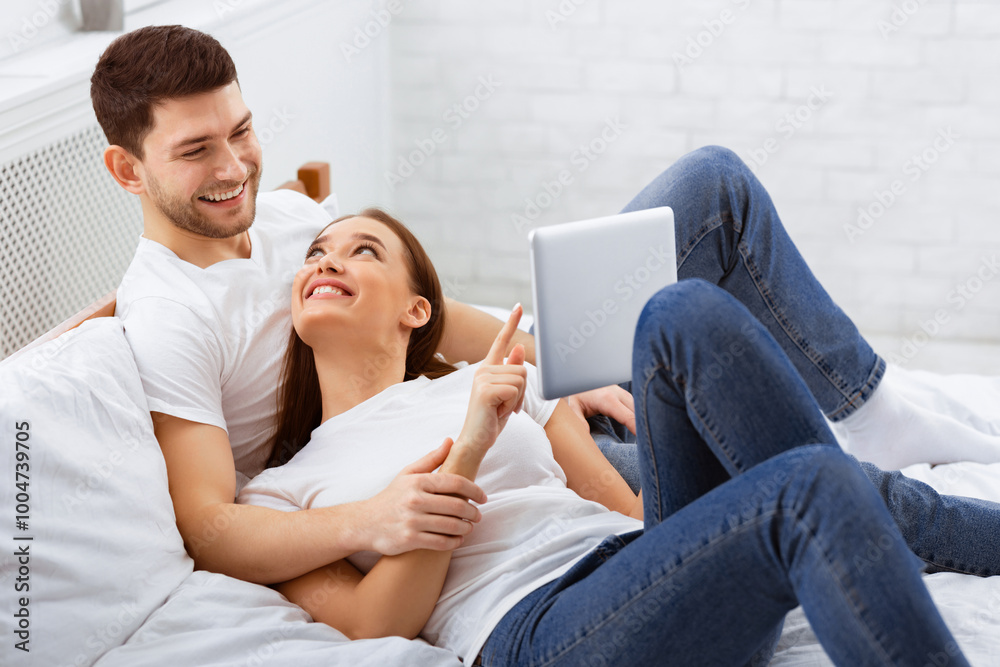 Cheerful Spouses Using Tablet Computer Lying In Bed At Home Relaxing On Weekend Morning. Selective Focus Cheerful Spouses Using Tablet Computer Lying In Bed At Home Relaxing On Weekend Morning. Selective Focus