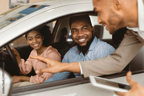 Joyful African Couple Buying New Car Talking With Salesman Testing Auto In Dealership Store. Selective Focus