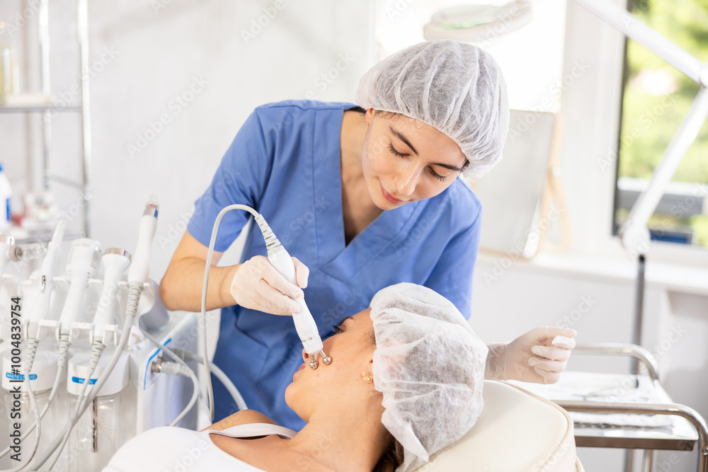 Obraz premium Young woman cosmetologist performs hardware facial microcurrent procedure to young female patient