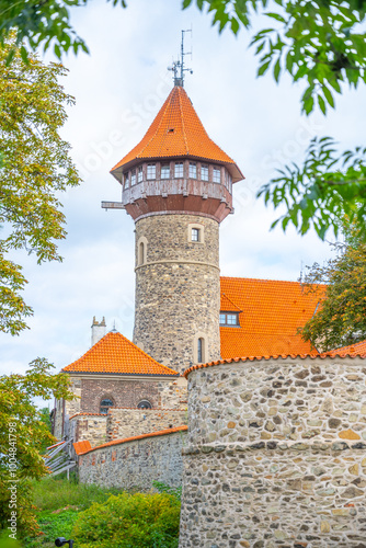 The lookout tower of Hnevin Castle stands tall against the sky, featuring distinctive red rooftops and stone walls, surrounded by lush greenery, showcasing its architectural beauty in Most, Czechia.