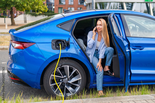 Young woman sits in a blue electric car, charging it while talking on her smartphone.