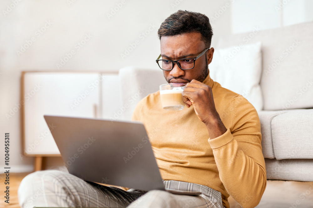 Serious African American Guy Reading News On Laptop And Drinking Coffee Sitting On Floor At Home. Selective Focus Serious African American Guy Reading News On Laptop And Drinking Coffee Sitting On Floor At Home. Selective Focus