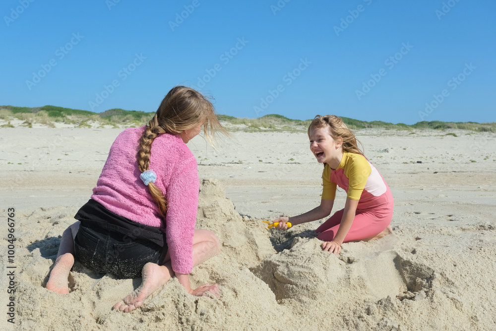 Two girls building a sandcastle on the beach under a clear blue sky. Two happy sisters having fun on the Atlantic ocean deserted beach Two girls building a sandcastle on the beach under a clear blue sky. Two happy sisters having fun on the Atlantic ocean deserted beach