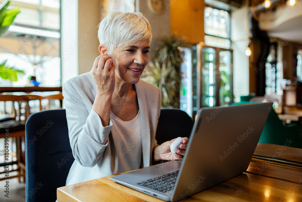Senior businesswoman working on laptop in a cafe with wireless earbuds Senior businesswoman working on laptop in a cafe with wireless earbuds