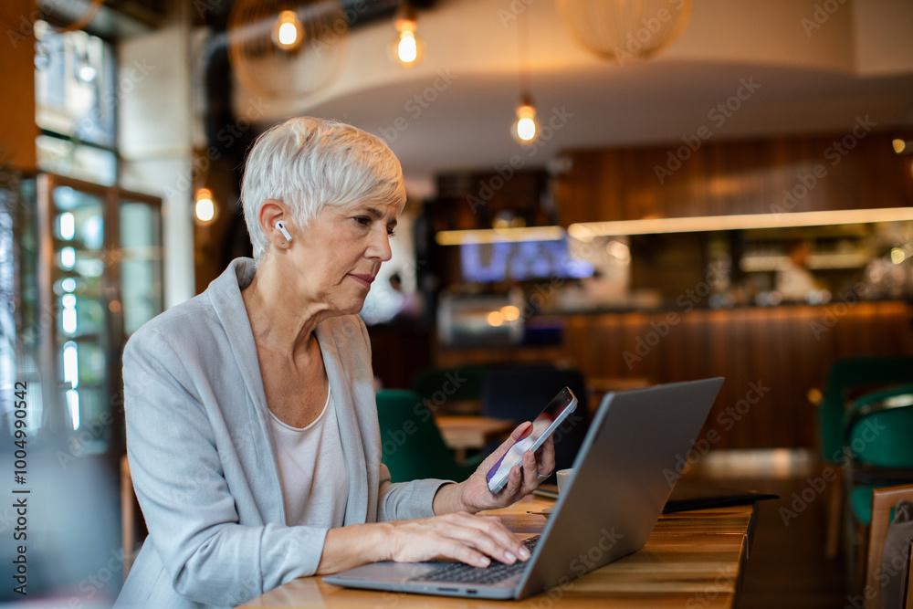 Senior businesswoman working on laptop in a cafe with wireless earbuds Senior businesswoman working on laptop in a cafe with wireless earbuds