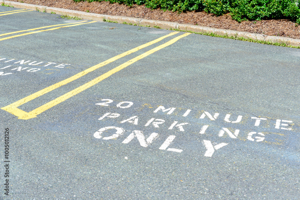 Obraz premium An asphalt parking lot with yellow double striped dividers. A white letter with 20-minute parking only painted on the ground. There's hedging and mulch as a border around the edge of the lot. 