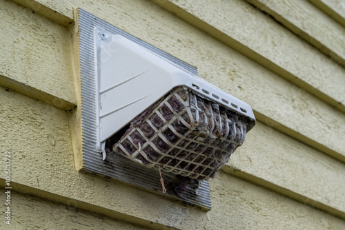 The exterior of a bright yellow house wall with a white dryer vent exposes the air vent which is clogged with hair, wood, and dirt in the mesh causing a fire hazard. The plastic unit guard is dirty.