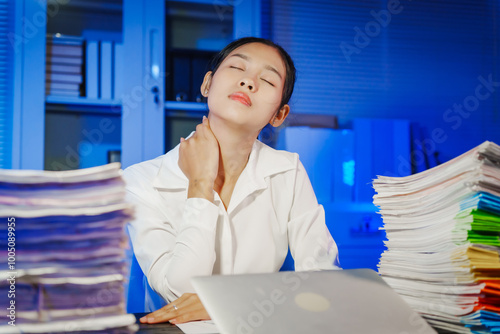 confident Asian businesswoman works late into the night, surrounded by a stack of papers in a busy office. Overtime leads to office syndrome symptoms like neck pain, headaches, and chronic stress. photo