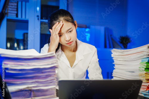 confident Asian businesswoman works late into the night, surrounded by a stack of papers in a busy office. Overtime leads to office syndrome symptoms like neck pain, headaches, and chronic stress. photo