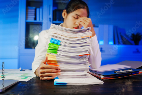 confident Asian businesswoman works late into the night, surrounded by a stack of papers in a busy office. Overtime leads to office syndrome symptoms like neck pain, headaches, and chronic stress. photo