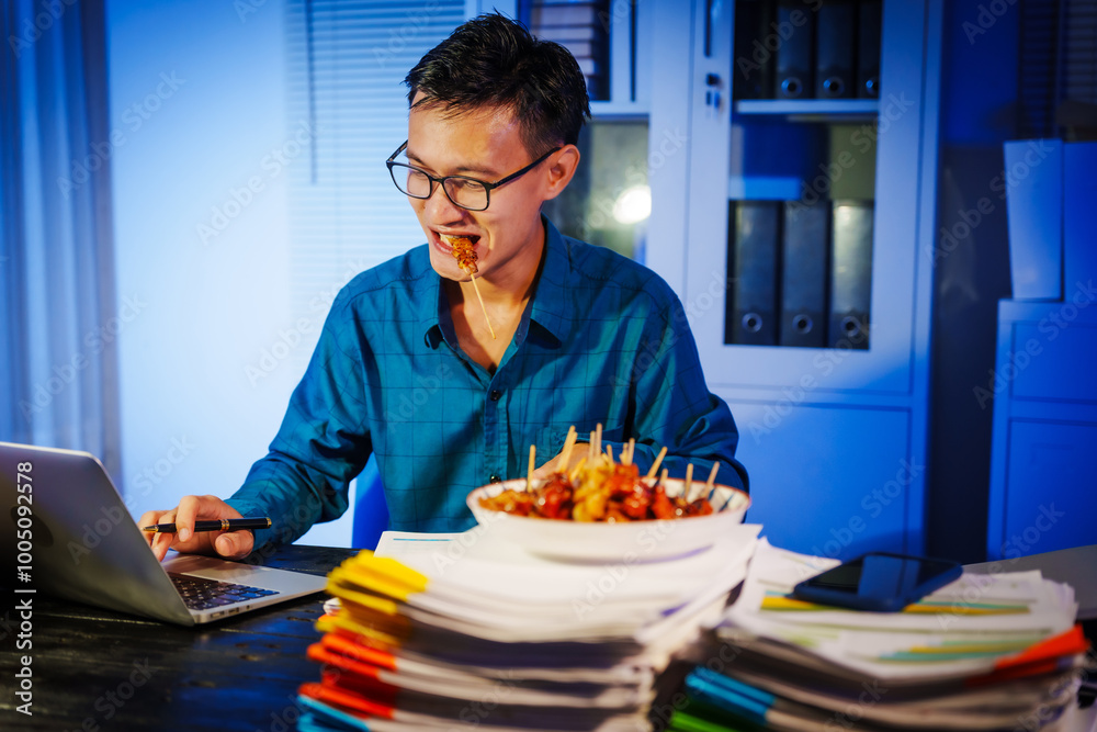 Asian businessman works late into the night, focused on a stack of papers on his black wooden desk. The office is dimly lit with a blue glow, reflecting the busy atmosphere of overtime. Asian businessman works late into the night, focused on a stack of papers on his black wooden desk. The office is dimly lit with a blue glow, reflecting the busy atmosphere of overtime.