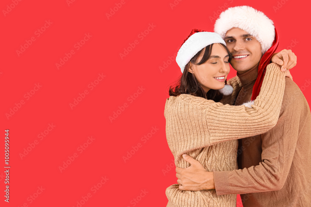 Happy young couple in Santa hats hugging on red background Happy young couple in Santa hats hugging on red background