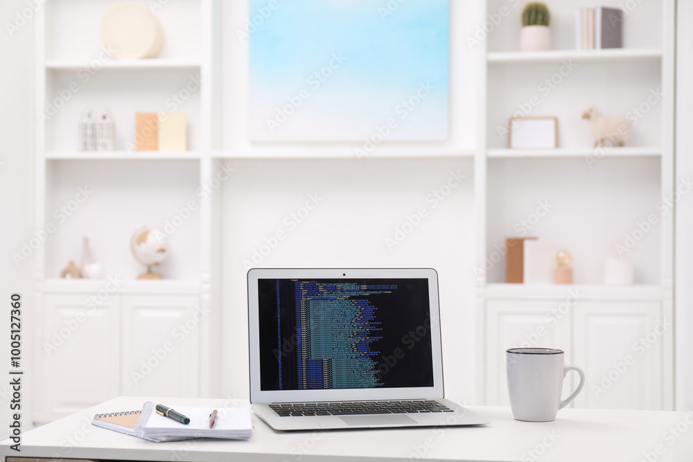 Programmer's workplace. Modern laptop with unfinished code, cup and stationery on white desk indoors