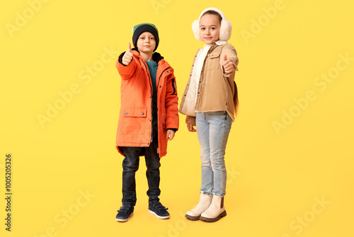 Cute little happy children in warm winter clothes showing thumbs-up gesture on yellow background