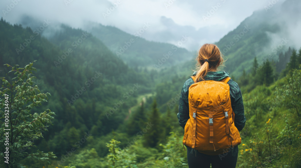 Naklejka premium woman with yellow backpack stands in lush green valley, surrounded by misty mountains. serene landscape evokes sense of adventure and tranquility