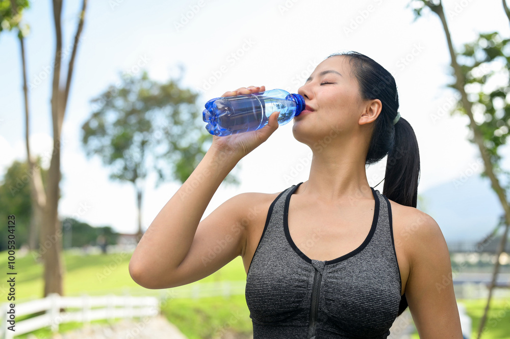 A fit woman in sportswear drinks from a water bottle, quenching her thirst after jogging in a park.
