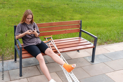 Tableau sur toile Teenage patient with a leg cast resting on a park bench, leveraging her smartphone for social interaction and video calls during her rehabilitation journey in the peaceful summer setting