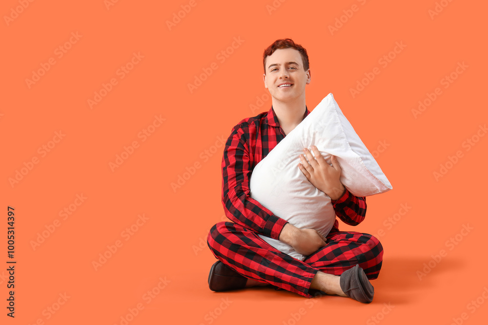 Fototapeta premium Handsome young happy man in checkered pajamas with pillow sitting on orange background
