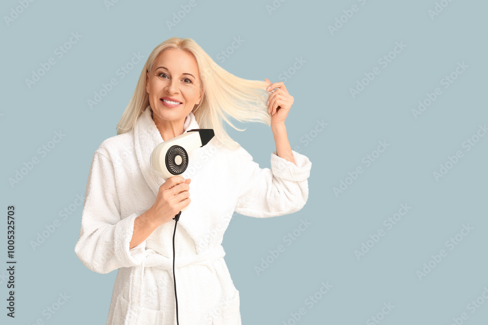 Fototapeta premium Mature woman in bathrobe with hair dryer on color background