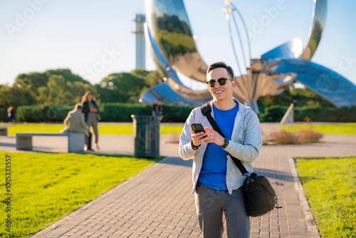 Young Latino man smiling and using his phone in front of the Floralis Genérica sculpture in Buenos Aires, Argentina, on a sunny day. photo