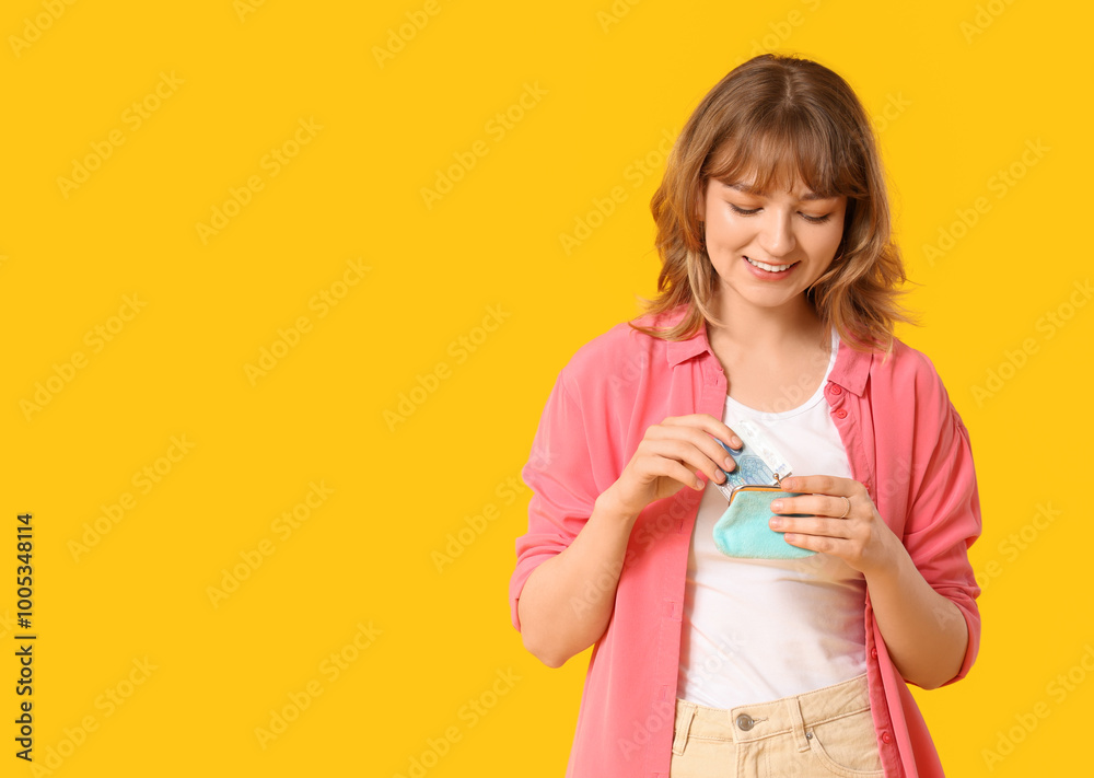Beautiful young woman putting money in blue purse on yellow background Beautiful young woman putting money in blue purse on yellow background