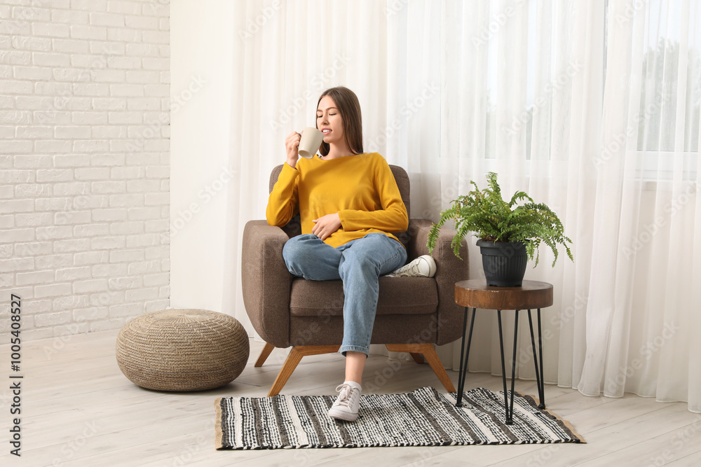 Beautiful young woman with mug in comfortable armchair near window in living room Beautiful young woman with mug in comfortable armchair near window in living room