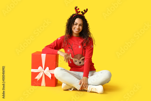 Happy young African-American woman in Christmas sweater and deer horns with gift box on yellow background