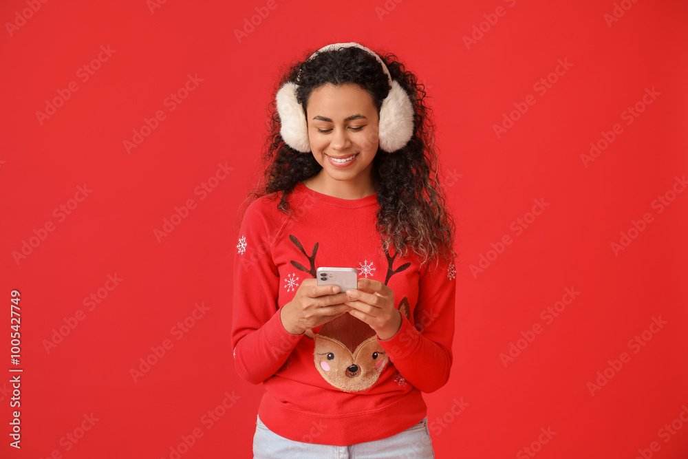 Beautiful young African-American woman in Christmas sweater with mobile phone on red background Beautiful young African-American woman in Christmas sweater with mobile phone on red background