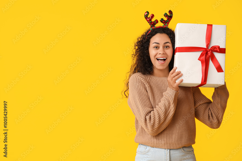 Happy young African-American woman in reindeer horns with Christmas gift box on yellow background Happy young African-American woman in reindeer horns with Christmas gift box on yellow background