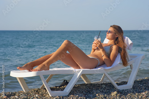 A young woman relaxes on a beach lounge chair by the ocean, sipping a refreshing drink under the warm sun. The serene coastal scenery and clear blue water capture the essence of a perfect summer day.
