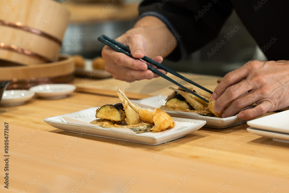 Obraz premium Japanese chef carefully plating tempura vegetables and seafood using chopsticks, creative presenting the dish on a rectangular ceramic plate in a kitchen setting