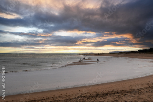 A beach with a cloudy sky and a few people walking on the sand