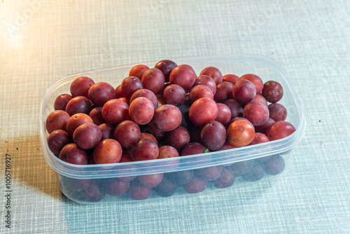 Heap of red tkemali fruits in transparent plastic container on light table. Prunus cerasifera, cherry plum, myrobalan plum photo