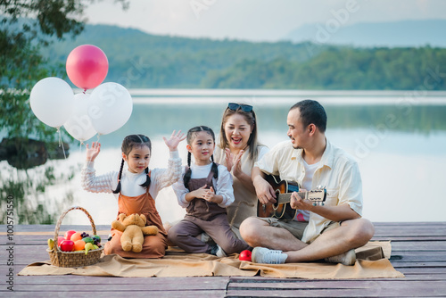 An Asian family sings and laughs happily on a wooden bridge by a lake, with the father playing the guitar.