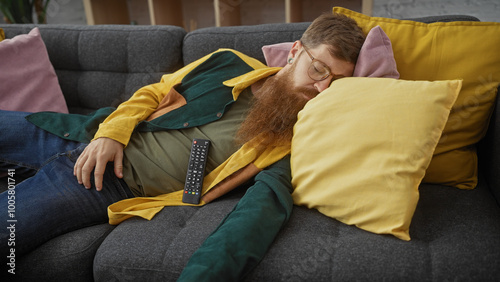 Bearded man sleeping on couch indoors with a remote control photo