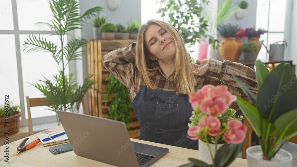 A young blonde woman stretches contently in her indoor flower shop surrounded by plants, showcasing a relaxed entrepreneur. A young blonde woman stretches contently in her indoor flower shop surrounded by plants, showcasing a relaxed entrepreneur.