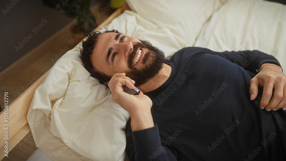 Smiling bearded man lying in bed talking on a smartphone at home Smiling bearded man lying in bed talking on a smartphone at home