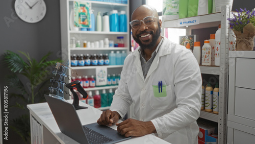 Smiling pharmacist, bald, man in a white coat working with a laptop in a modern pharmacy interior with shelves of products and medical supplies