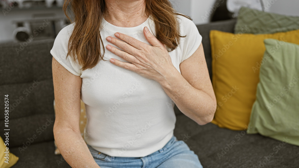 Fototapeta premium Mature woman experiencing chest discomfort sitting on a sofa indoors, touching her heart area with a concern expression.