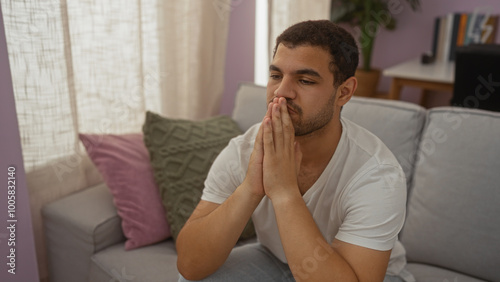 Thoughtful young hispanic man sitting indoors on a sofa in a living room, contemplating with hands clasped, creating a serene and introspective atmosphere in a cozy home setting.