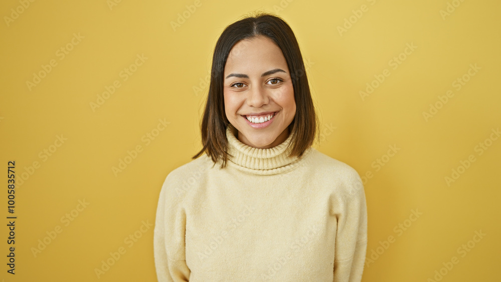 A cheerful young hispanic woman with a beautiful smile poses in an ivory sweater against a yellow isolated background.