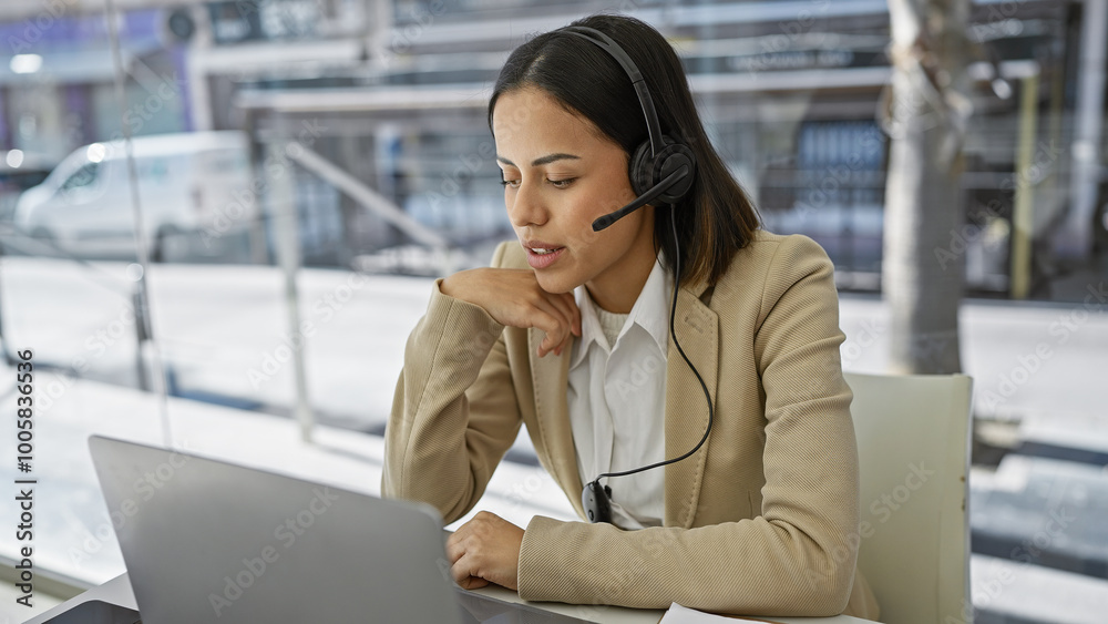 A professional young woman with a headset concentrates on her laptop in a bright office setting.