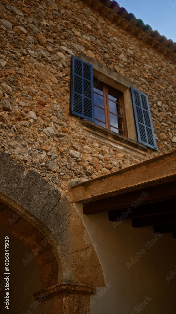 Stone building facade with arched entrance and blue shutters on a window under a bright sky Stone building facade with arched entrance and blue shutters on a window under a bright sky