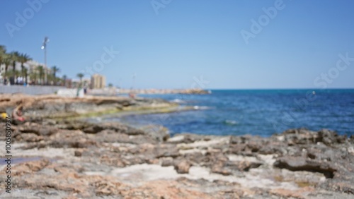 Blurry coastal scene featuring rocky shore, blue ocean, and distant people enjoying sunny weather along a promenade in an outdoor setting